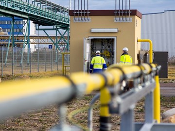 Wasserstoffprojekt HYPOS: H2-Netz im Vordergrund befinden sich Rohrleitungen, im Hintergrund Männer mit Arbeitskleidung in einer technischen Anlage