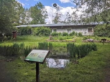 Naturförderungsvereinigung im Vordergrund ist ein Teich auf einer Wiese mit einer Beschreibungstafel, im Hintergrund ein grauer Flachbau